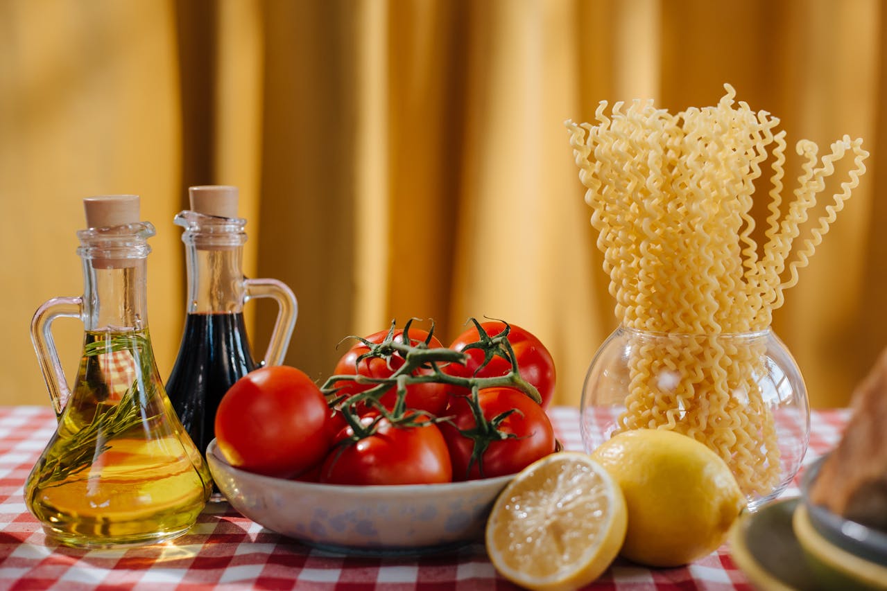 Botellas de aceite de oliva virgen extra y vinagre balsámico junto a un cuenco con tomates frescos, pasta y limones en una mesa de cocina, listos para preparar recetas mediterráneas.